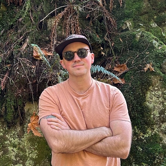 Jared Andres, wearing a cap and round sunglasses and a peach t-shirt, arms crossed, standing in front of a mossy rock wall.