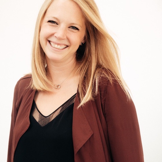 Katie Pelletier, smiling, with shoulder-length blond hair, wearing a maroon jacket over a black top.